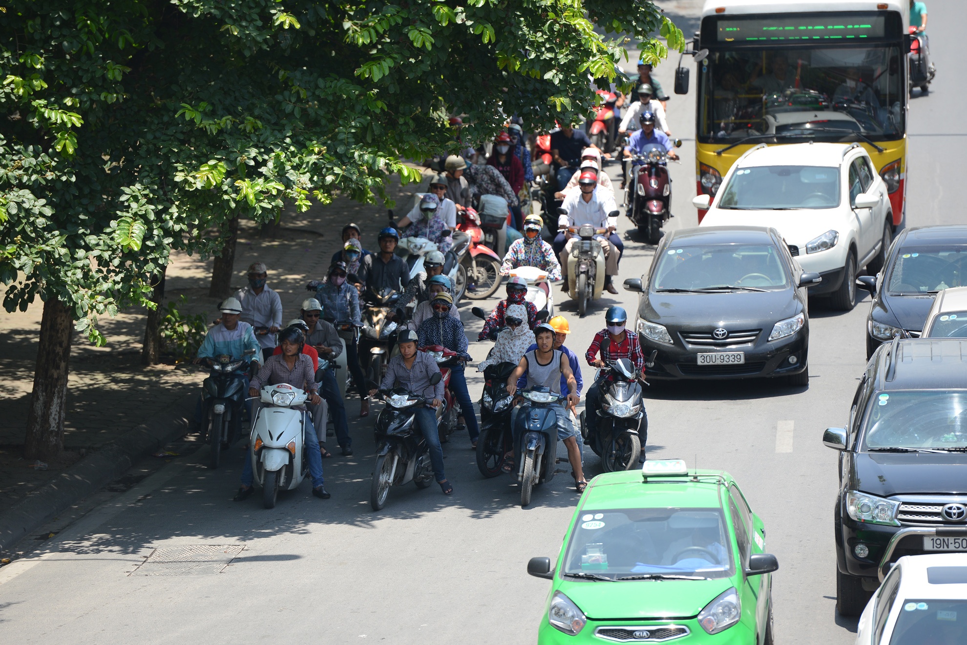Người đi xe máy dừng tại nơi có bóng cây chờ đèn tín hiệu.