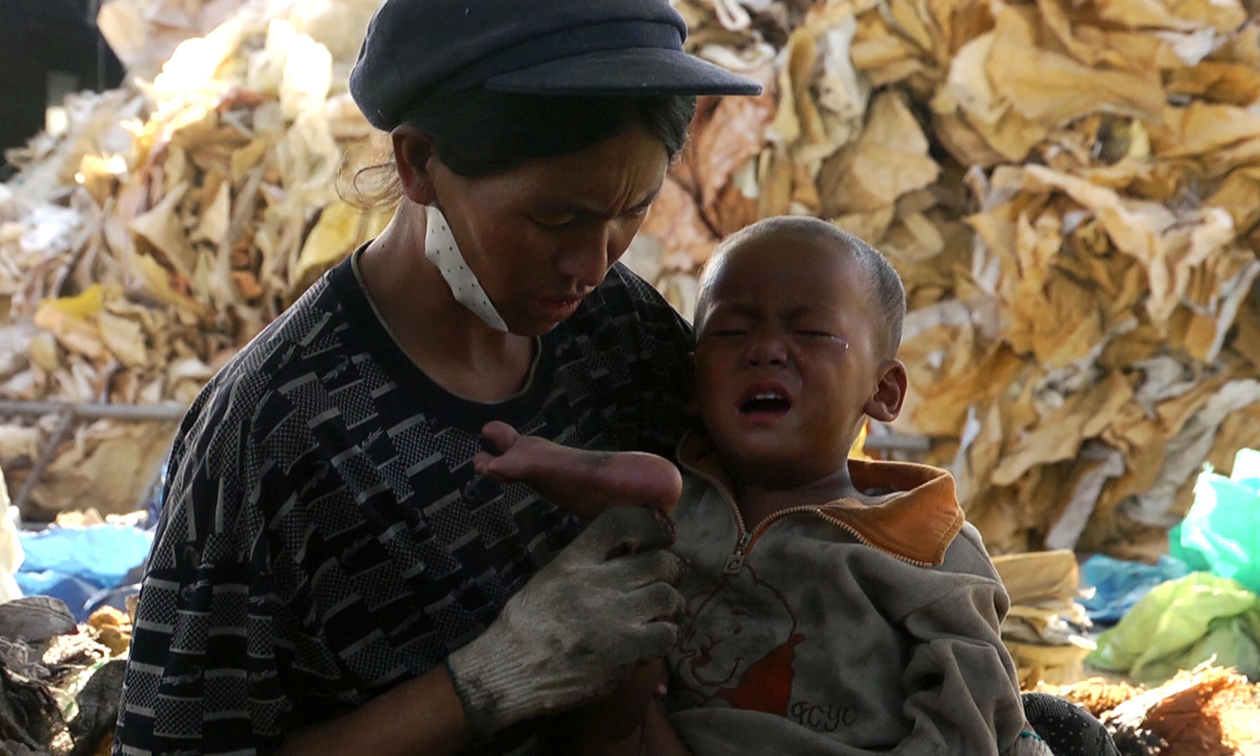 Một người phụ nữ đang vỗ về con trai, sau khi cậu bé giẫm phải một vật sắc trong xưởng xử lý rác. Những mảnh vật thể nhọn vương vãi rất nhiều trong các xưởng, đe dọa công nhân và trẻ em tại đây.