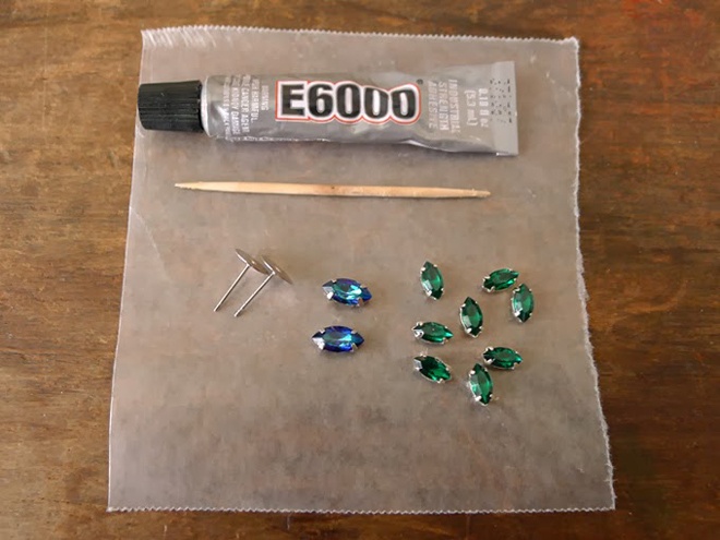 Đầu tiên, hãy sưu tầm đầy đủ các nguyên vật liệu sau:  1 ống keo E600, tăm và giấy sáp (hoặc 1 tấm bìa nhựa trong), 8 viên đá crystal màu xanh lục bảo và 2 viên đá crystal màu xanh corban (có thể chọn màu tùy theo sở thích của bạn), 2 miếng đế hoa tai.