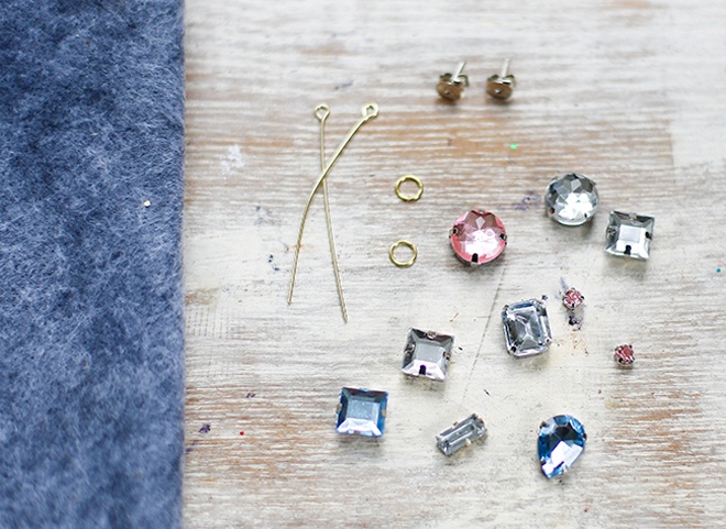 Để bắt tay vào chế tạo kiểu hoa tai độc đáo này, bạn cần chuẩn bị các nguyên vật liệu sau: từ 5 – 10 hạt đá crystal hoặc hạt nhựa giả đá với kích thước, kiểu dáng và màu sắc khác nhau, 2 chiếc đế khuyên tai, 2 vòng tròn nhỏ (hoặc nhẫn đeo tay loại nhỏ) mạ vàng, 2 cây kim mạ vàng hoặc dùng kềm bẻ cong 2 sợi dây đồng mạ vàng thành 2 đầu tròn nhỏ như hình minh họa và 1 tấm vải nỉ màu xanh nhạt, kéo và keo dán.