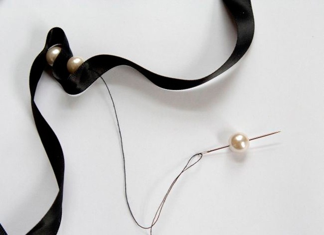 Đầu tiên, dùng kéo cắt sợi dây chuyền để lấy ngọc trai ra. Luồn chỉ đen qua khoảng 1/3 sợi dây ruy băng rồi kết 1 hạt ngọc trai vào. Gấp 1 phần dây ruy băng lên mặt trên của hạt ngọc trai và tiếp tục khâu chỉ luồn qua sợi dây ruy băng như hình minh họa.