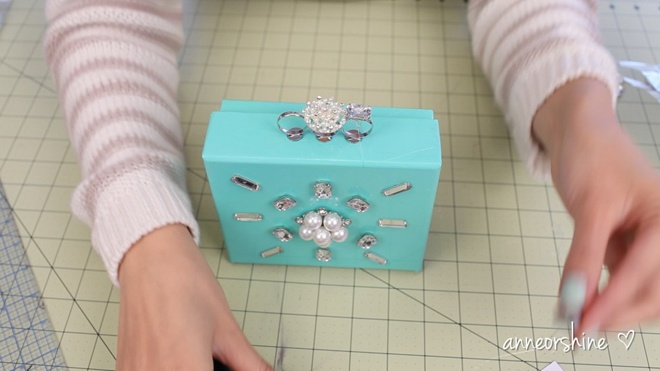 Gắn 2 chiếc nhẫn vào cạnh bên của clutch, xen giữa là các chi tiết trang trí khác như chùm hoa bằng ngọc trai giả, hạt đá to … để tạo thành phần quai cầm.