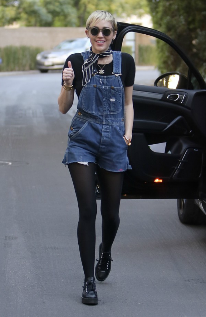 Miley Cyrus kết hợp một chiếc quần short bò yếm với áo phông đen, khăn quàng nhỏ, vòng cổ Chanel, giày thể thao và quần tất đen dày.