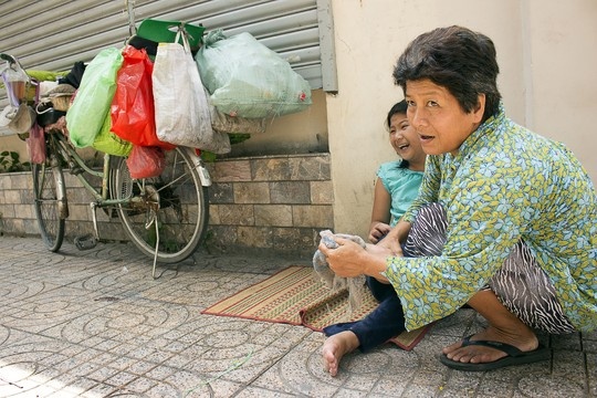 Trải một chiếc chiếu bên lề đường, bà Mai chăm sóc con gái lớn Khưu Thị Ái My (12 tuổi) bị bại não bẩm sinh trong lúc đợi Huỳnh Giao tan học. Trước đây, bà thường tranh thủ thời gian này, vừa cõng Ái My vừa đi lượm ve chai, nhặt rác. Nhưng gần đây Ái My thường xuyên lên cơn động kinh, bà chỉ có thể ngồi một chỗ chăm sóc con.
