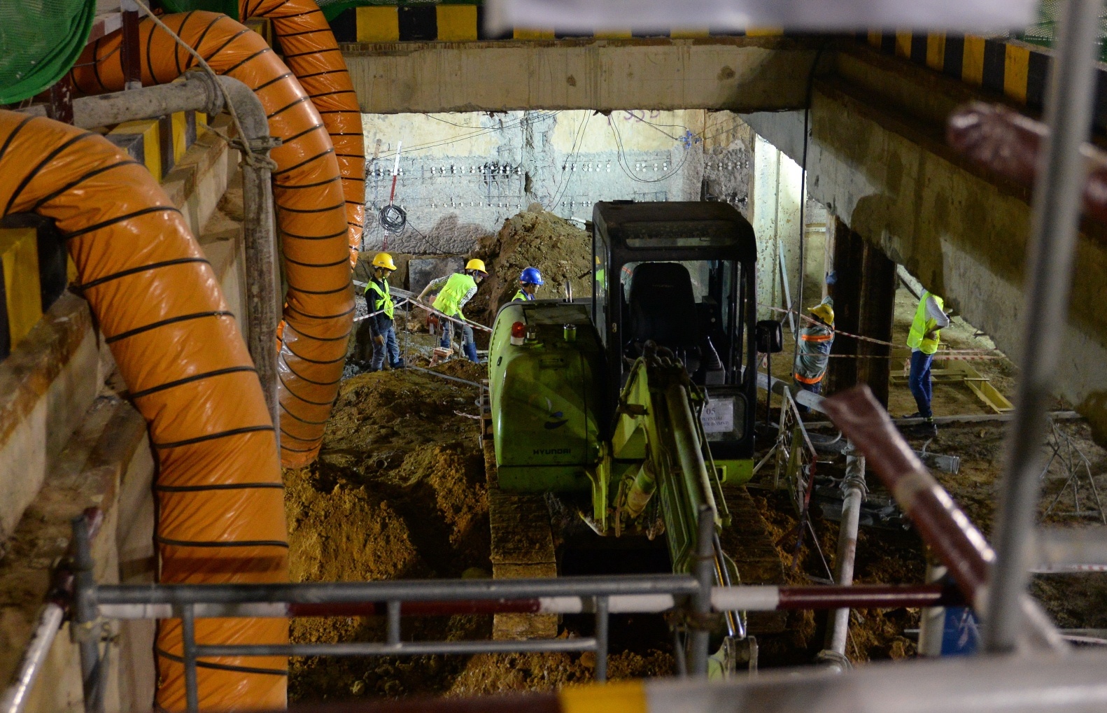 Trên tầng hai của văn phòng công trình làm bằng thùng xe container, năm kỹ thuật viên đang ngồi xử lý số liệu quan trắc do các kỹ thuật viên khác đi đo đạc ở các tòa nhà gần đó đưa về. Kỹ thuật viên Âu Tấn Hậu nói rằng công trình này còn sử dụng thiết bị quan trắc tự động của Đức và Singapore, được gắn trên hai tòa nhà cao trong khu vực và truyền dữ liệu về cho kỹ thuật viên 24 lần mỗi ngày. Trong ảnh: Một máy xúc đất đậu sẵn dưới tầng ngầm để đưa đất ra ngoài.