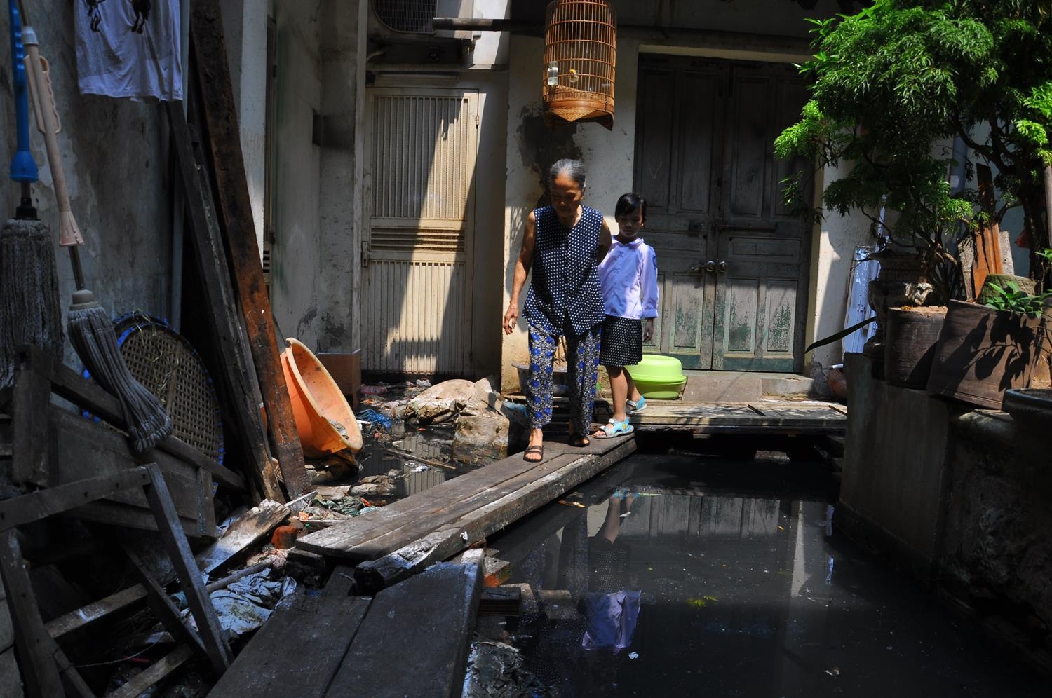 Dù tuổi cao sức yếu nhưng hàng ngày bà Phạm Thị Điểm (80 tuổi) phải đưa cháu gái tên An (6 tuổi) đi qua cây cầu khỉ ngập nước thải để tới lớp. Vì mắt không còn minh mẫn, bà Điểm và cháu gái đã từng nhiều lần sẩy chân ngã xuống nước bẩn. Bà thì bị thương ở chân, còn cháu gái thì phải về thay quần áo và bị muộn học. Ngoài ra, mọi sinh hoạt của 7 người trong gia đình gồm 3 thế hệ của bà Điểm đều duy trì khép kín trong ngôi nhà cũ.