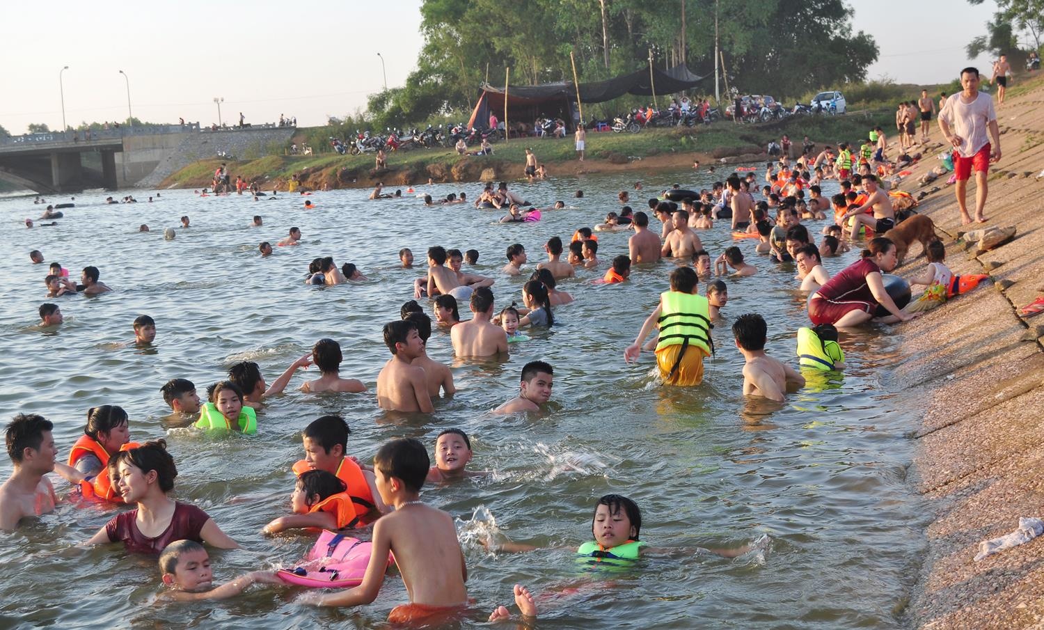 Dọc bờ đập khoảng 500m, một hàng dài những chiếc xe máy đã chen nhau xếp hàng để xe. Cứ vài phút lại có tốp gần chục chiếc xe chở hết công suất. Người ôm phao bế cháu nhỏ xuống đập nước. Bên dưới đập, hàng trăm người đủ các lứa tuổi bơi và nghịch nước. Những thanh niên 15 tuổi trở lên tự tin bơi ra giữa đập, nơi mực nước sâu nhất. Còn những phụ huynh đưa con nhỏ và gia đình đi tắm chỉ ở gần bờ. 