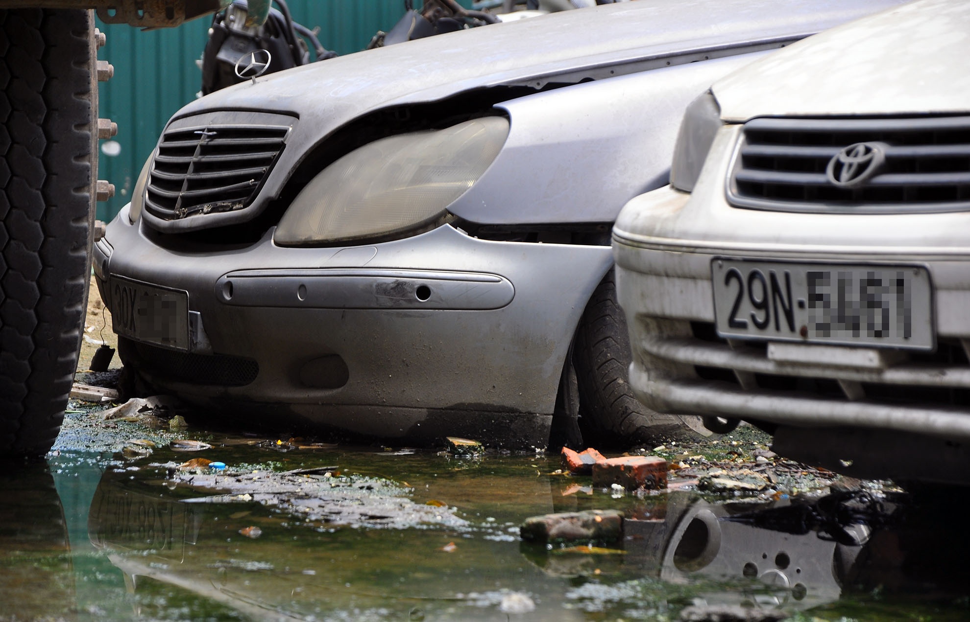 Một chiếc Meredes đời cũ đắt tiền ngập bùn trong bãi giữ phương tiện vi phạm. Bốn bánh xe này đã bị xì hơi, ba-đờ-sốc bong tróc.