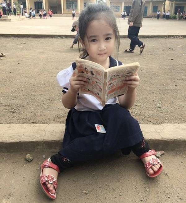 Bé gái xinh xắn như búp bê được cô giáo chụp ảnh trong lúc đang ngồi đọc truyện tranh trong sân trường.