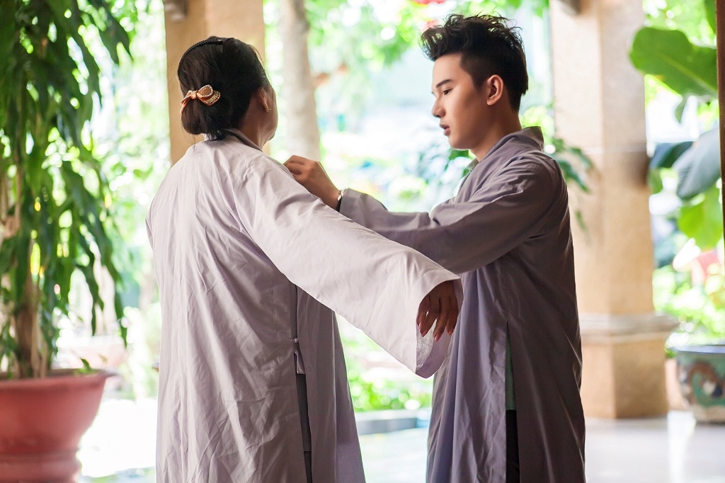 Khác với hình ảnh lung linh thường thấy trên sân khấu, Khắc Minh cùng mẹ giản dị đến viếng chùa trong bộ áo tràng màu lam. Trước khi vào bên trong chùa, cậu em út của nhóm Ayor còn tự tay mặc áo tràng cho mẹ. 