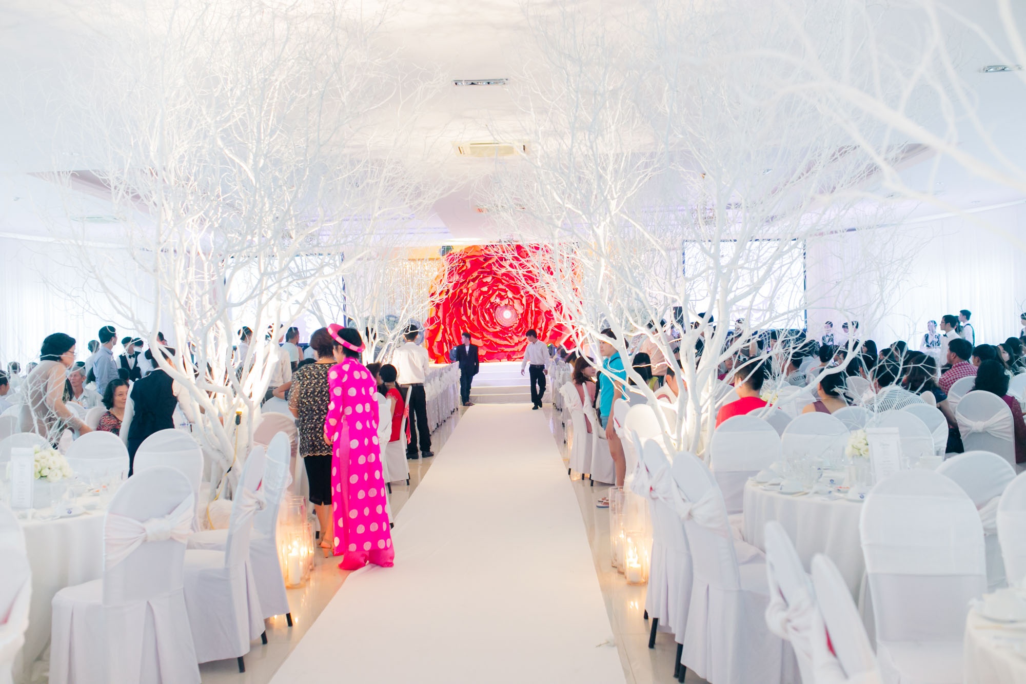 Toàn cảnh không gian tiệc cưới của Công Vinh và Thủy Tiên trước giờ G. Không gian cưới bao phủ màu trắng - màu sắc yêu thích của cô dâu Thủy Tiên. Điểm nhấn trong phòng tiệc chính là bông hồng đỏ khồng lồ đặt giữa trung tâm của sân khấu. Bông hồng này được làm thủ công trong 2 ngày.