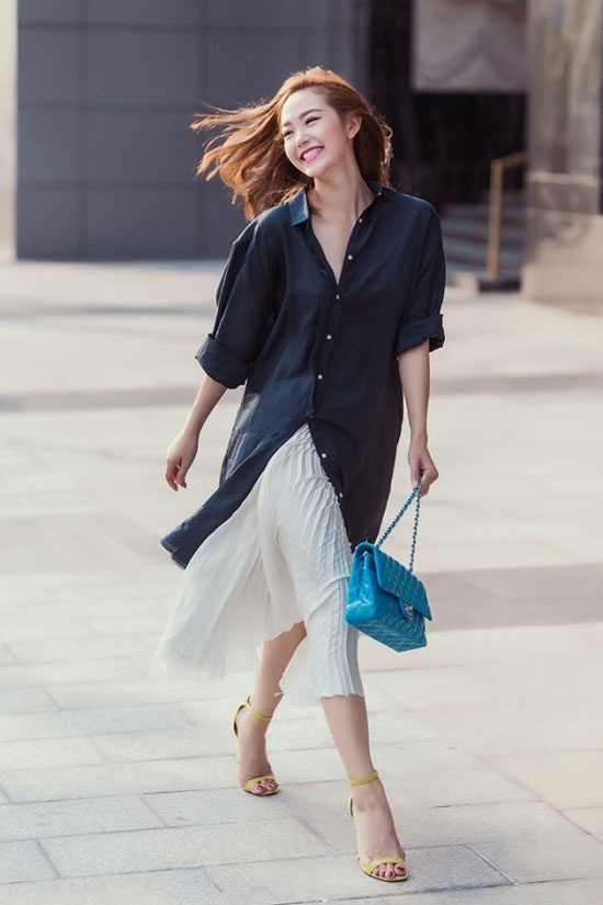 Minh Hằng xinh tươi và rạng rỡ trên phố với áo sơ mi đen dáng rộng kết hợp chân váy xếp ly trắng. Phụ kiện nổi bật của túi đeo màu xanh, xăng đan vàng khiến tổng thể set đồ rất trẻ trung và có sức hút.