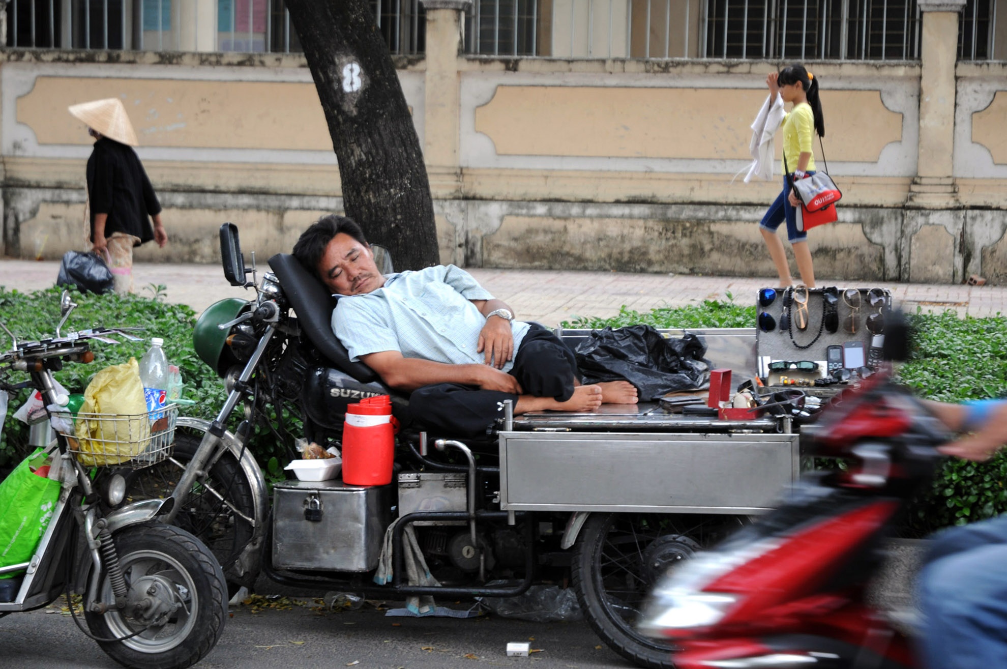 Một người đàn ông khuyết tật buôn bán đồ cũ, tranh thủ ngủ trưa dù đường phố đông xe.