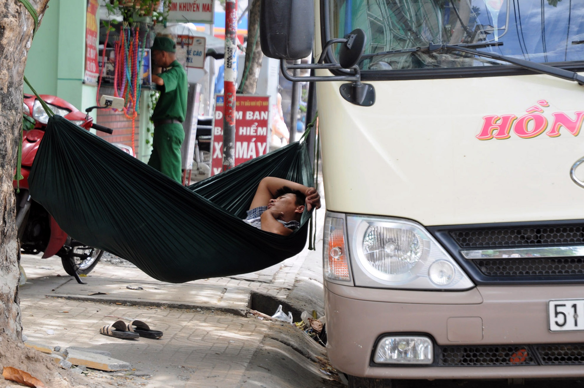 Một bác tài xế xe khách đang ngủ trưa trên chiếc võng mắc vội để chờ đón khách.