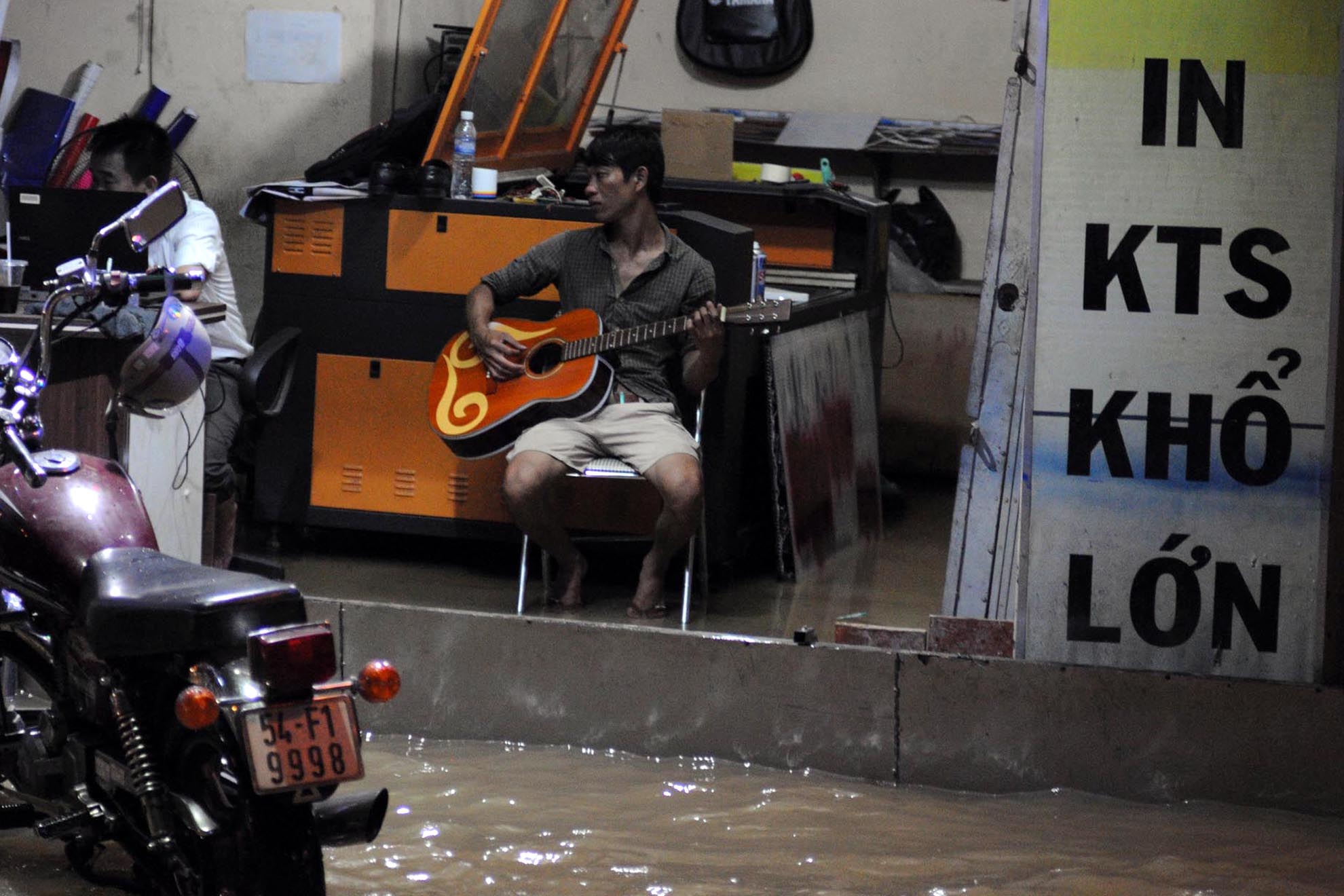 Chàng trai ngán ngẩm ngồi ôm đàn ngồi cảm thán trong khi nước ngập hết cửa hàng in sao, photocopy mà không thể làm gì được.